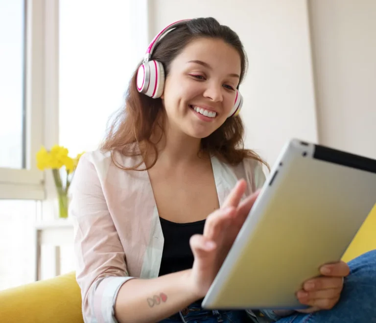 Mujer joven sonriente usando auriculares y una tablet en un ambiente acogedor, mientras interactúa con la pantalla y disfruta de su tiempo libre.